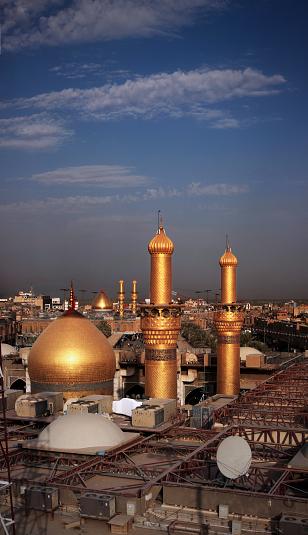 Golden dome and two golden minarets of a Karbala shrine at dusk, with a second golden dome in the distance