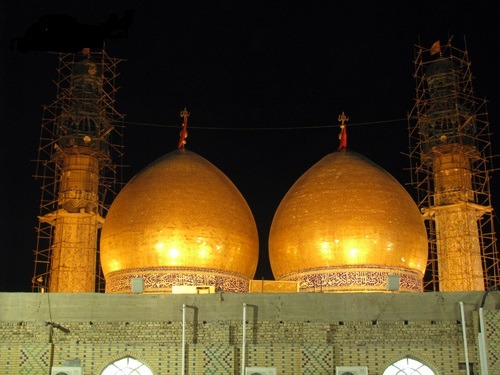 Twin golden domes lit at night, flanked by two minarets under scaffolding