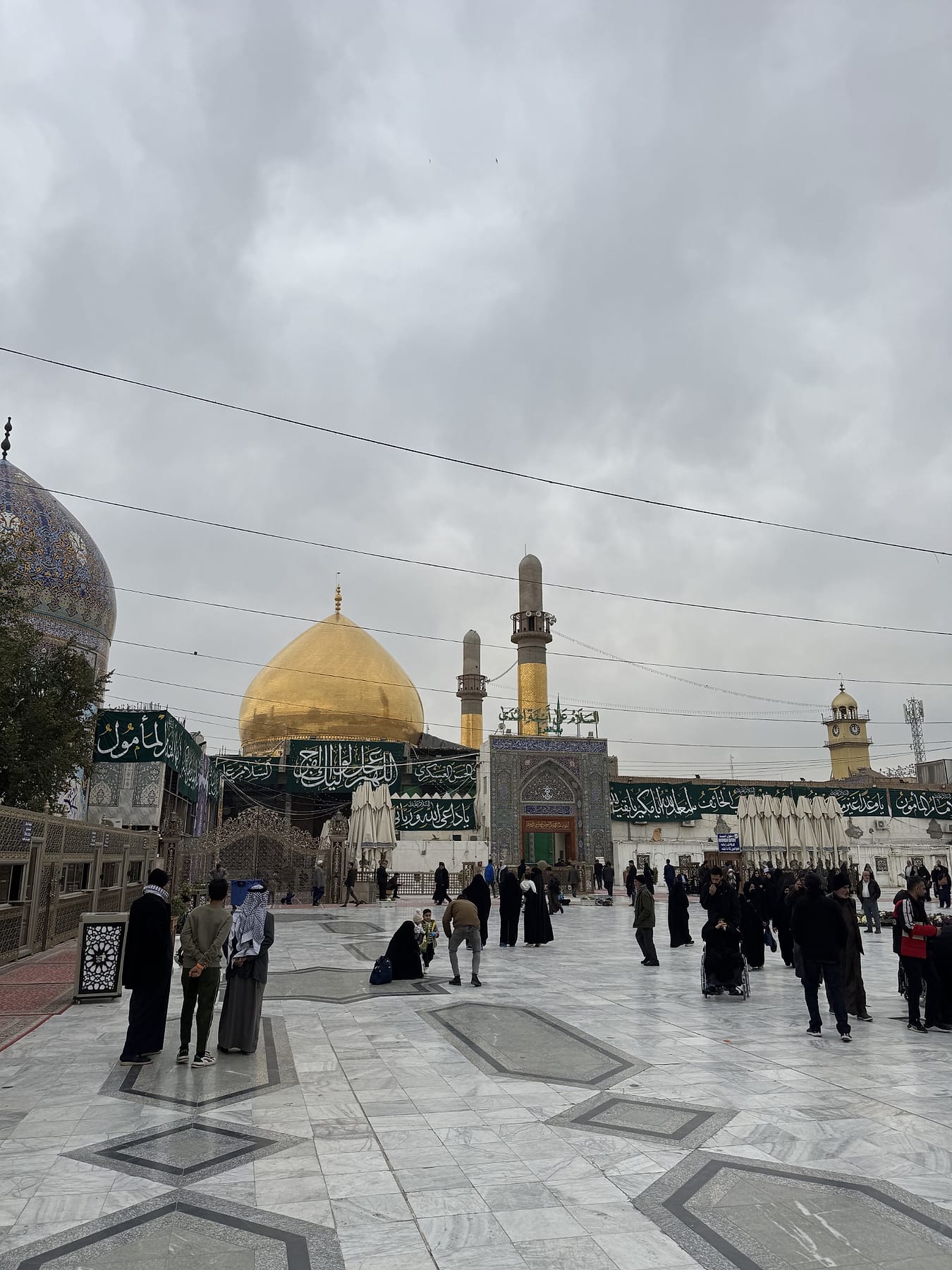 A large golden dome and two minarets above the shrine walls, with visitors crossing a marble courtyard under a cloudy sky