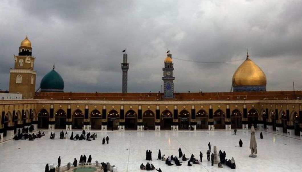 A wide white marble courtyard under a cloudy sky, ringed by arcades, with a golden dome, a green dome and minarets beyond; small groups of visitors sit on the floor