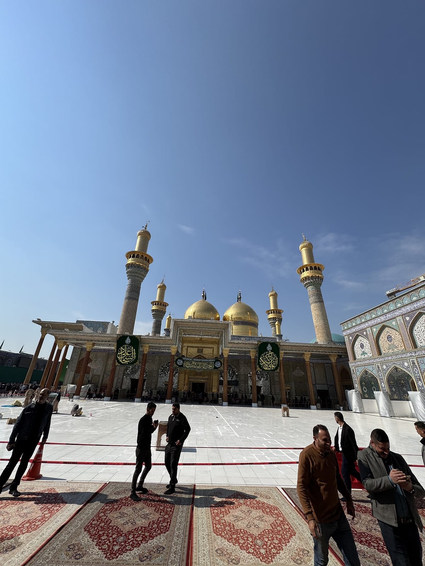 Golden domes and minarets above the shrine walls by day, with visitors in the courtyard