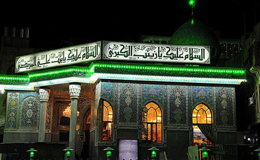 A small tiled shrine building at night, lit in green, with a green dome behind and Arabic calligraphy greeting Lady Zaynab above the entrance